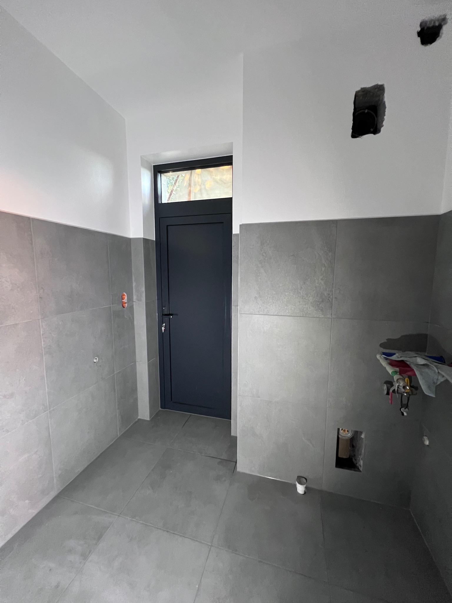 Szara łazienka w trakcie remontu, widoczne płytki na ścianach i podłodze, otwory w ścianach na instalacje, ciemne drzwi w tle.