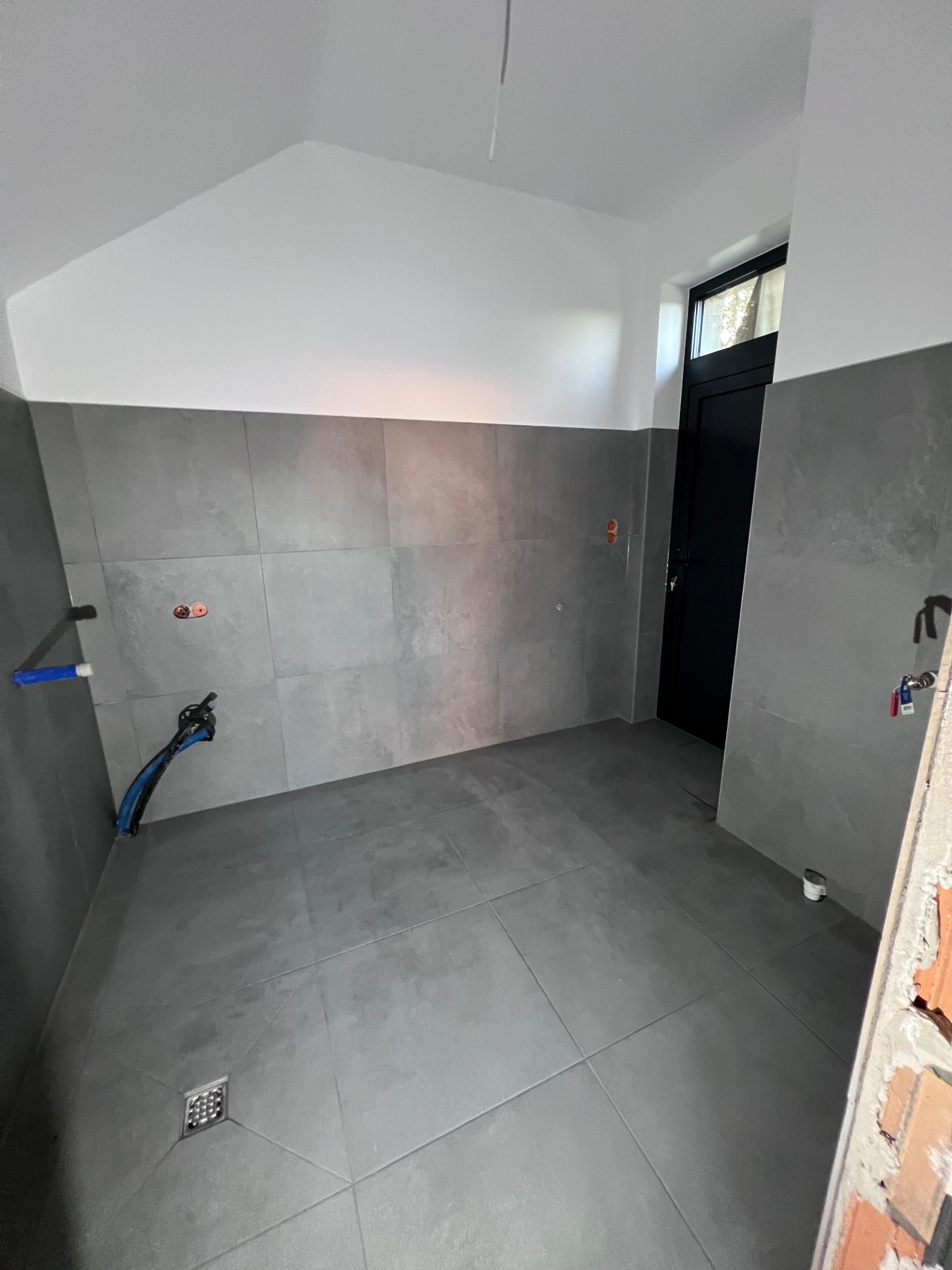 Wykończona łazienka w szarościach z dużymi płytkami, widoczny odpływ liniowy, przygotowane instalacje wodne i elektryczne, czarne drzwi wejściowe.