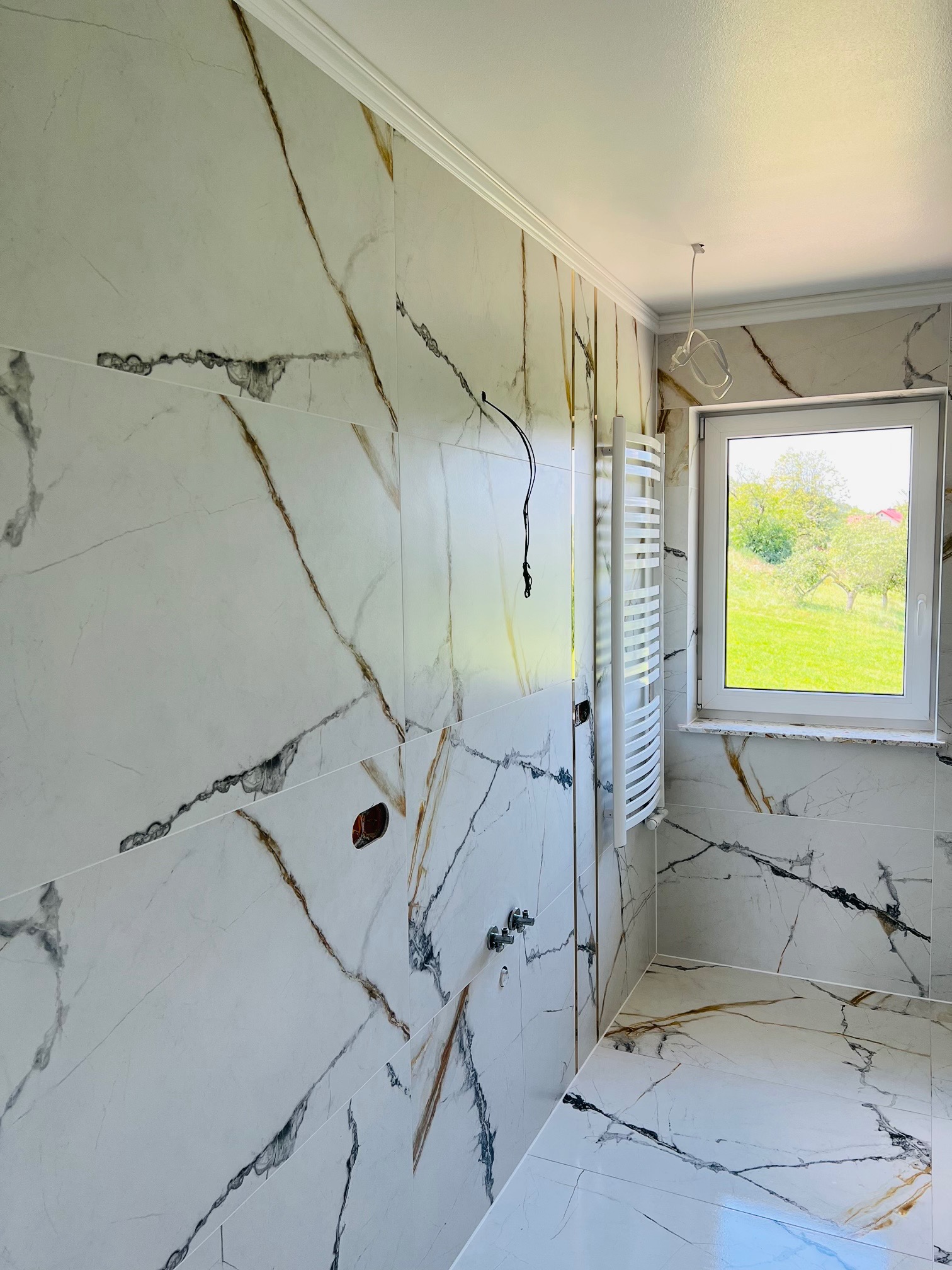 Łazienka w trakcie wykańczania: ściany i podłoga wyłożone białymi płytkami imitującymi marmur z wyraźnymi brązowymi i czarnymi żyłami, widoczny grzejnik drabinkowy, okno z widokiem na zielony ogród...