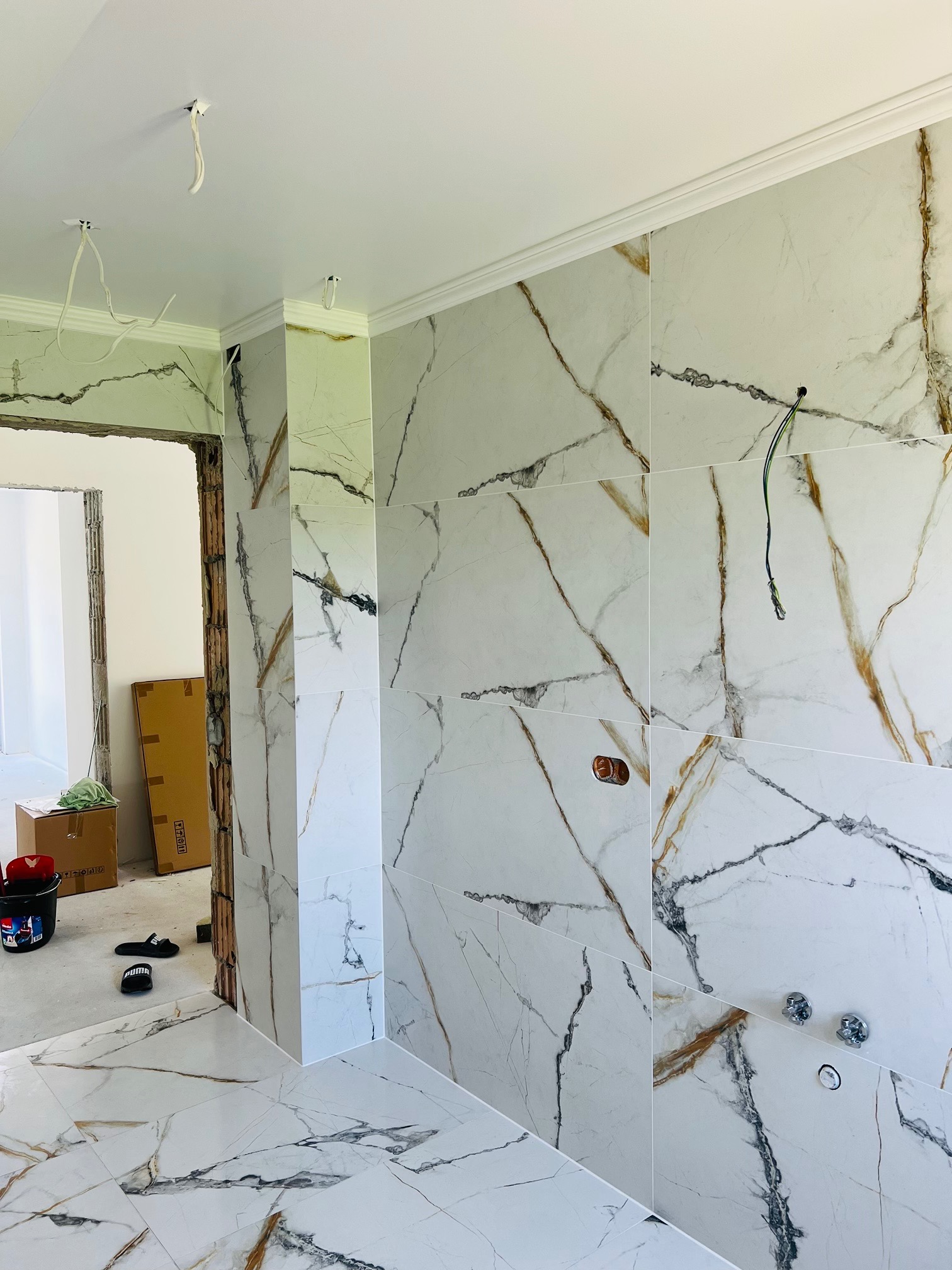 Wnętrze łazienki w trakcie wykańczania, z ułożonymi białymi płytkami imitującymi marmur na ścianach i podłodze, widoczne wystające przewody elektryczne, otwory na armaturę, oraz fragmenty...