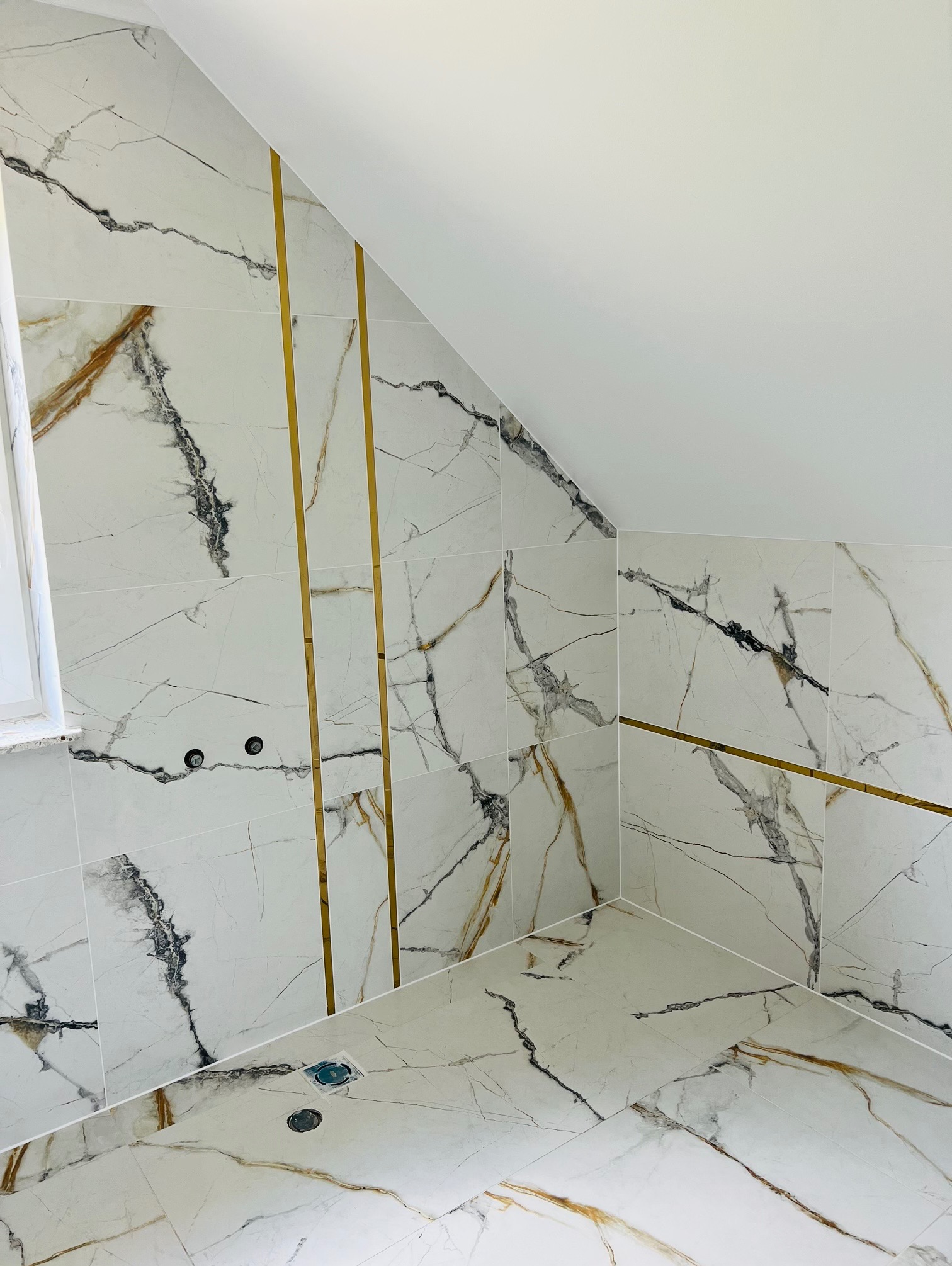 Wykończona łazienka na poddaszu z białymi płytkami imitującymi marmur z złotymi wstawkami, widoczny odpływ liniowy i wystające przyłącza wodne.