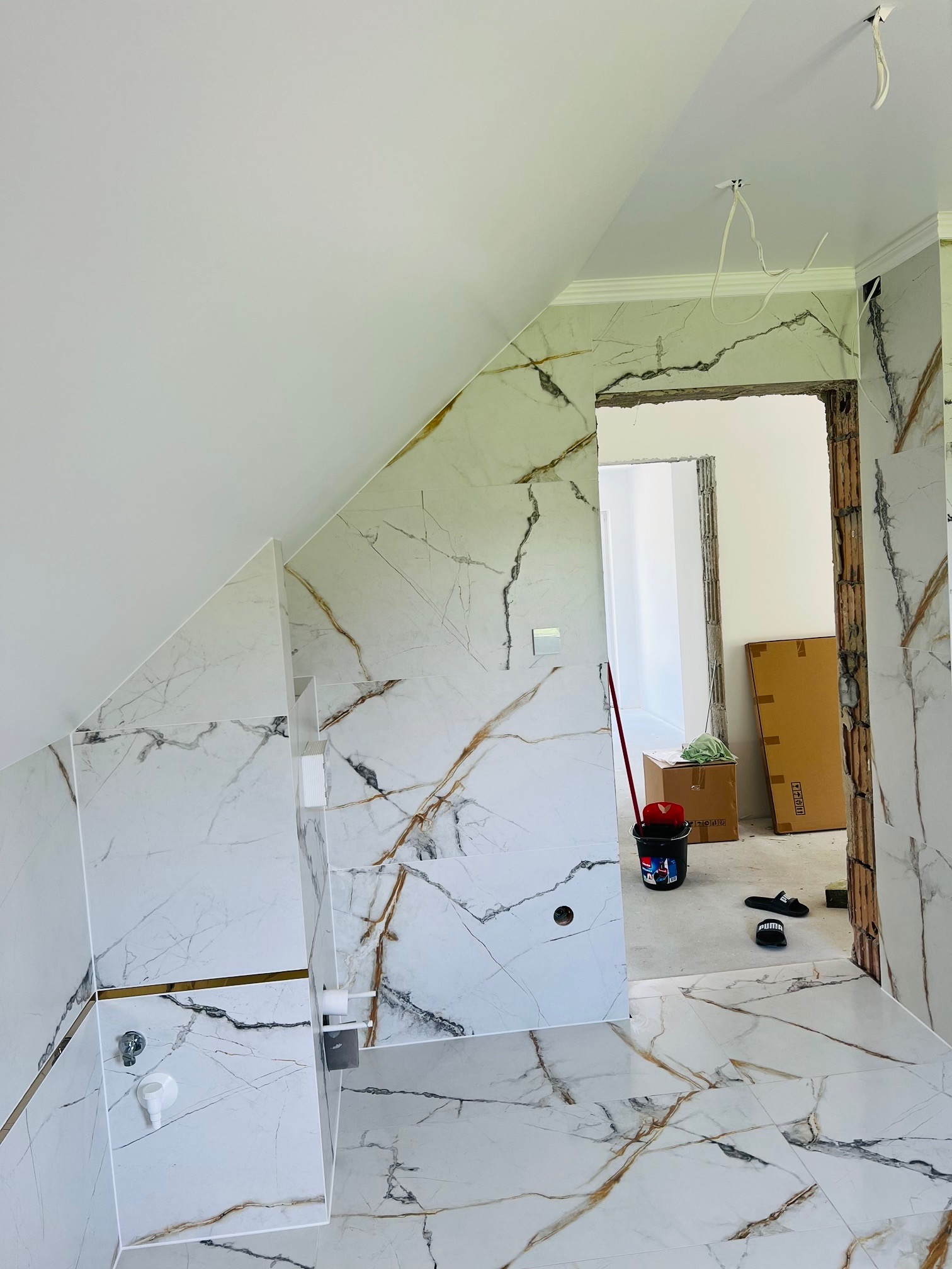 Łazienka w trakcie remontu z ułożonymi białymi płytkami imitującymi marmur z złotymi żyłami, widoczne wiadro, kartony i niedokończone futryny drzwiowe.