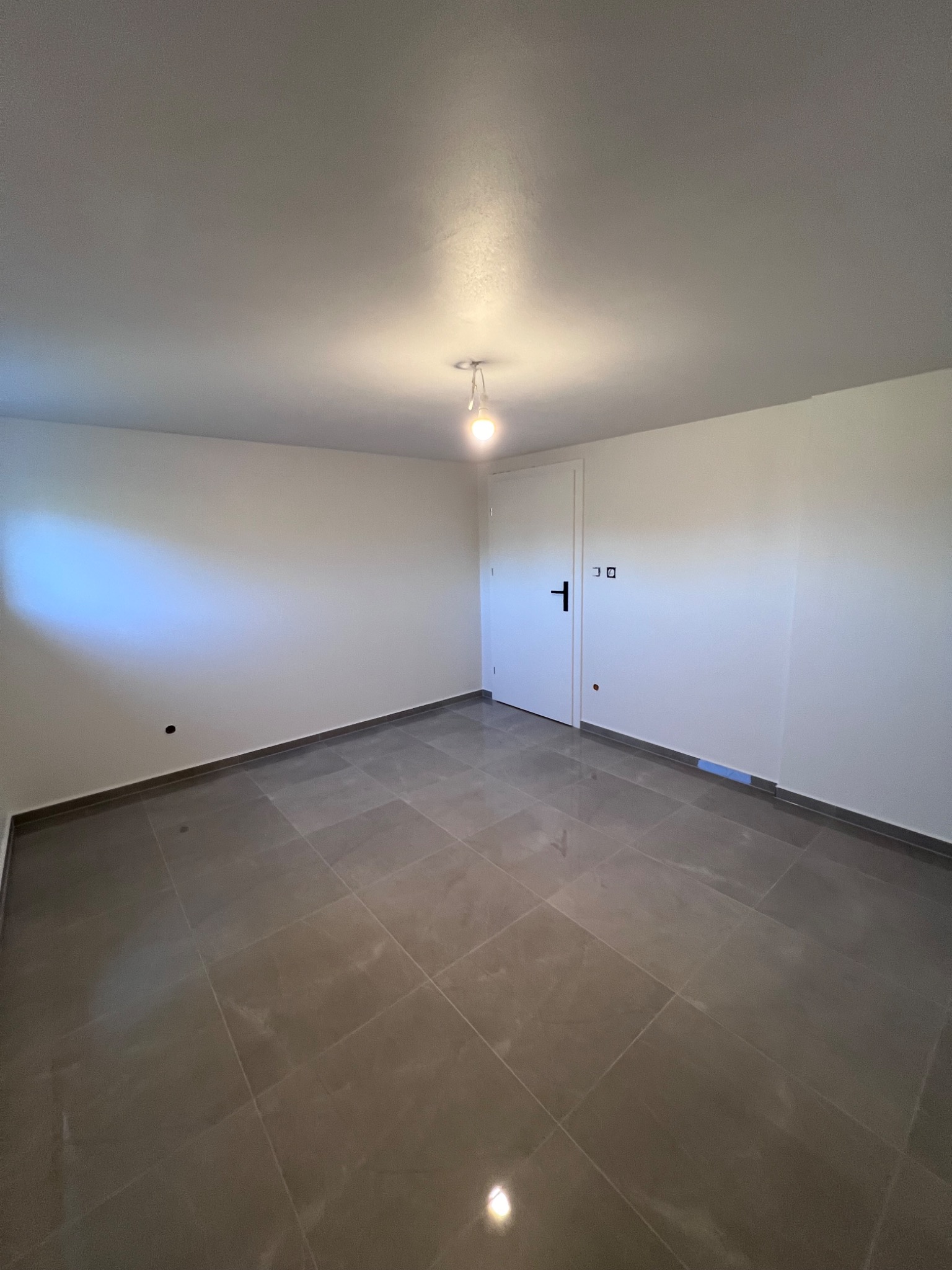 Puste pomieszczenie z szarymi płytkami podłogowymi ułożonymi w kwadraty, białe ściany, białe drzwi z czarną klamką i żarówka zwisająca z sufitu.