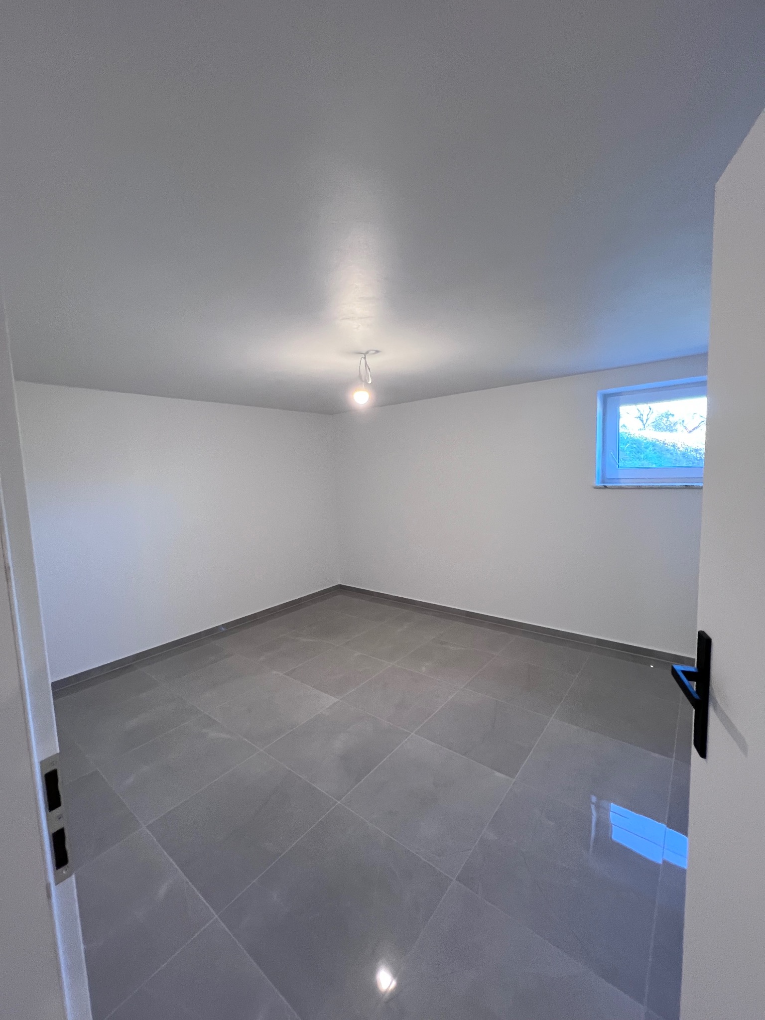 Puste, wykafelkowane pomieszczenie z szarymi, kwadratowymi płytkami podłogowymi, białymi ścianami, pojedynczą żarówką i oknem z widokiem na zieleń, widziane z perspektywy otwartych drzwi.