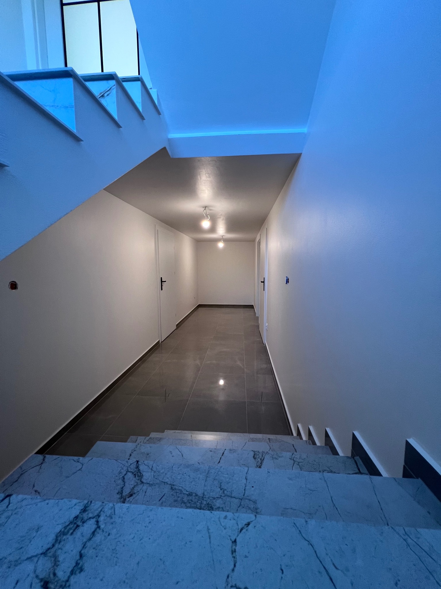 Korytarz w nowym budynku z marmurowymi schodami, szarą podłogą z płytek, pomalowanymi na biało ścianami i oświetleniem LED.
