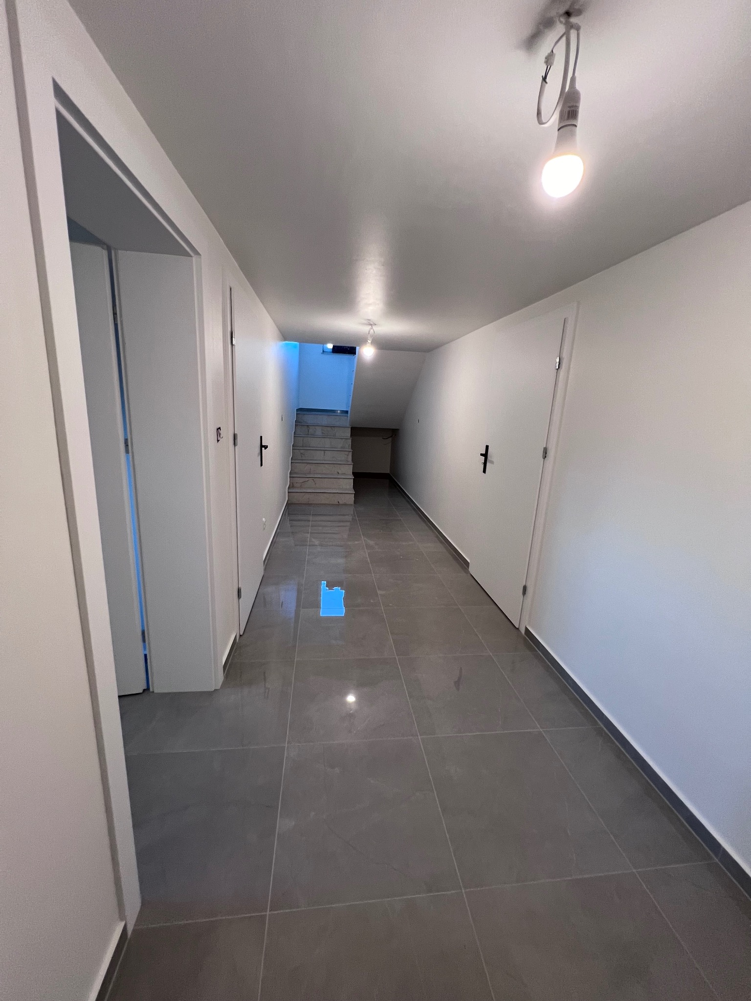 Długi korytarz z szarymi płytkami podłogowymi, białymi ścianami i drzwiami po obu stronach, oświetlony żarówkami LED, prowadzący do schodów z błękitnym światłem w tle.