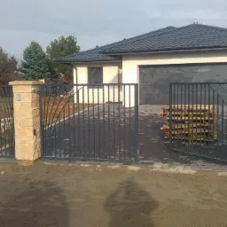 Nowoczesna, metalowa brama wjazdowa z dekoracyjnymi elementami, osadzona na murowanych słupkach z piaskowca, na tle domu z szarą dachówką i garażem, widoczne palety z kostką brukową na podjeździe.