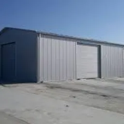 Szara hala przemysłowa z trzema bramami segmentowymi, widoczna z zewnątrz w słoneczny dzień, na betonowym placu, z zarysem drzew i budynków w tle.