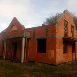 Surowy stan budynku jednorodzinnego z czerwonej cegły, widoczne otwory okienne i zarys wejścia, częściowo zadaszone, na tle jesiennego krajobrazu.