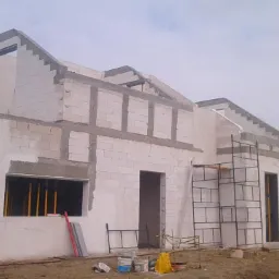 Budowa domu z białych bloczków, widoczne zbrojenia nad oknami i drzwiami, rusztowania przy ścianie bocznej, w tle dom z czerwonym dachem.