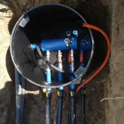 Niebieski moduł hydrauliczny z zaworami w czarnej studzience, zamontowany w wykopie ziemnym.