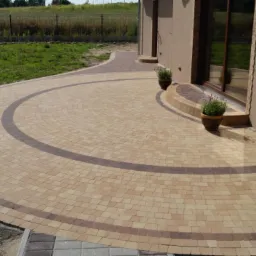 Półokrągły podjazd z beżowej kostki brukowej z ciemnobrązowymi akcentami, prowadzący do domu z doniczkami z lawendą przy wejściu, otoczony trawnikiem i ogrodzeniem.