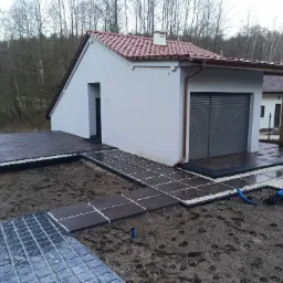 Nowoczesny taras z ciemnych płyt betonowych, obrzeża z jasnego grysu, widok na budynek z szarą elewacją i czerwonym dachem, w tle drzewa.