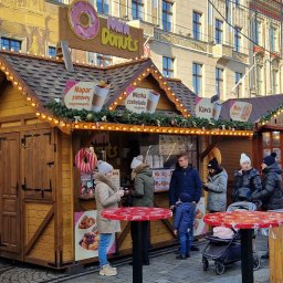 Domki Jamróz - Drewniany stragan z mini pączkami na jarmarku bożonarodzeniowym, oświetlony lampkami, z klientami i stolikami na pierwszym planie. Budynki w tle.