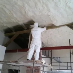 Pracownik w kombinezonie ochronnym aplikuje białą pianę na skośny sufit poddasza, stojąc na rusztowaniu. Widoczne drewniane elementy konstrukcyjne dachu i fragmenty ściany.