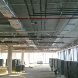 Wnętrze w stanie deweloperskim z widocznymi instalacjami pod sufitem: rury, przewody wentylacyjne w izolacji, kable elektryczne. Stosy płyt na paletach czekają na montaż przy oknach.