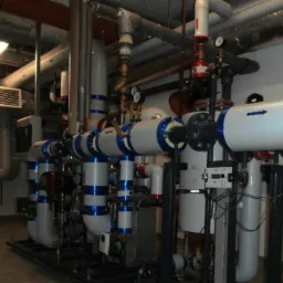 Złożona instalacja rur w kotłowni z manometrami, zaworami i izolacją w kolorach biało-niebieskich, widoczna także tabliczka informacyjna 'Sieć Cieplna'.