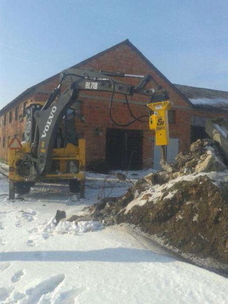 Żółty młot hydrauliczny zamontowany na ramieniu koparki Volvo BL71B, rozbijający gruz na śniegu, na tle ceglanego budynku gospodarczego.