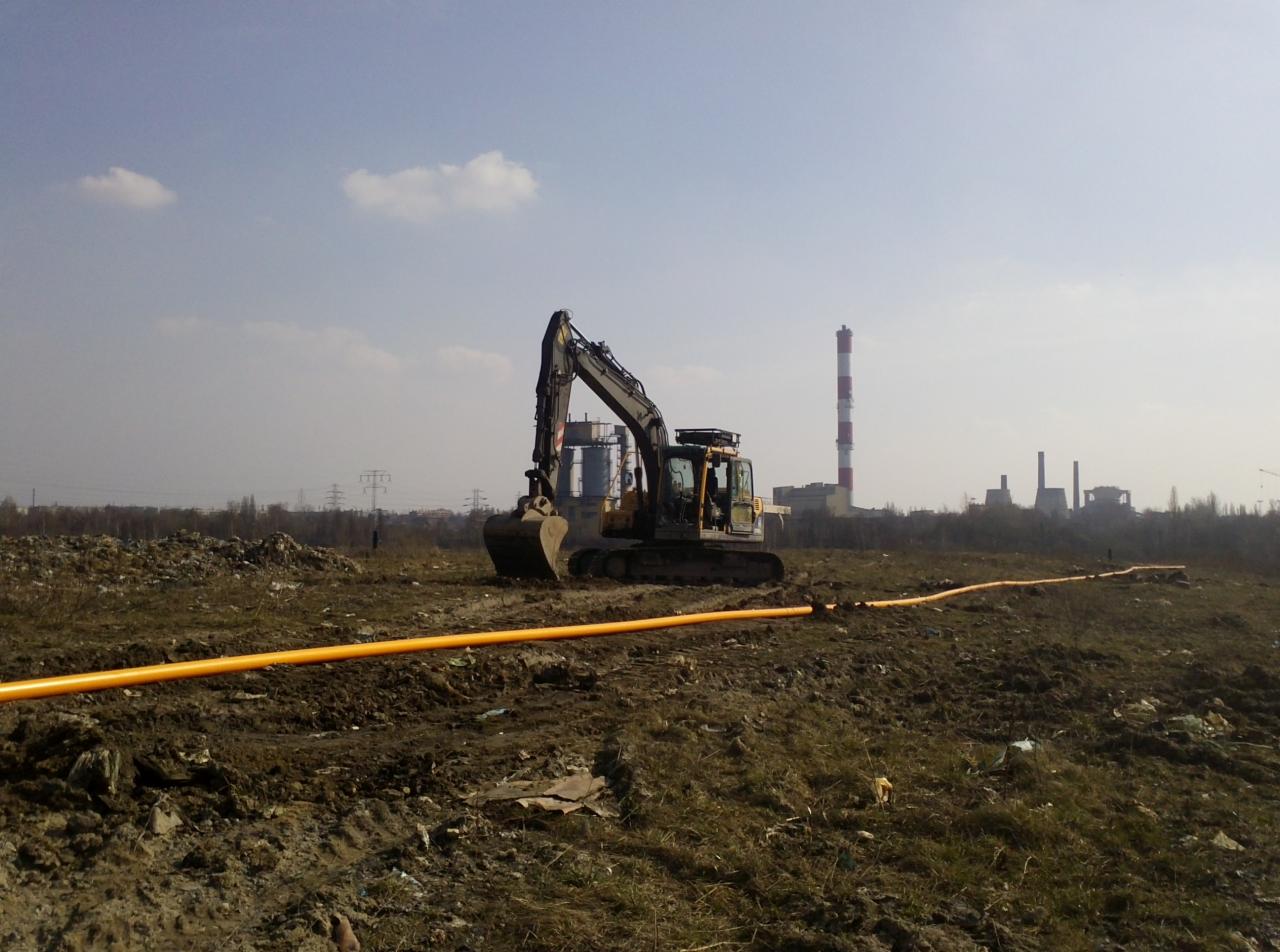 Żółty rurociąg ułożony w ziemi, na tle koparki i zabudowań przemysłowych z wysokim kominem.