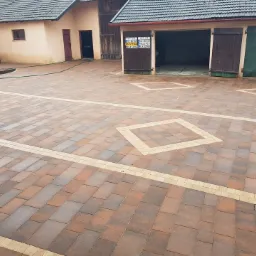 Ułożona kostka brukowa w odcieniach brązu i beżu, z dekoracyjnymi elementami w kształcie rombów i pasów, na tle budynków gospodarczych z otwartymi wrotami garażowymi.