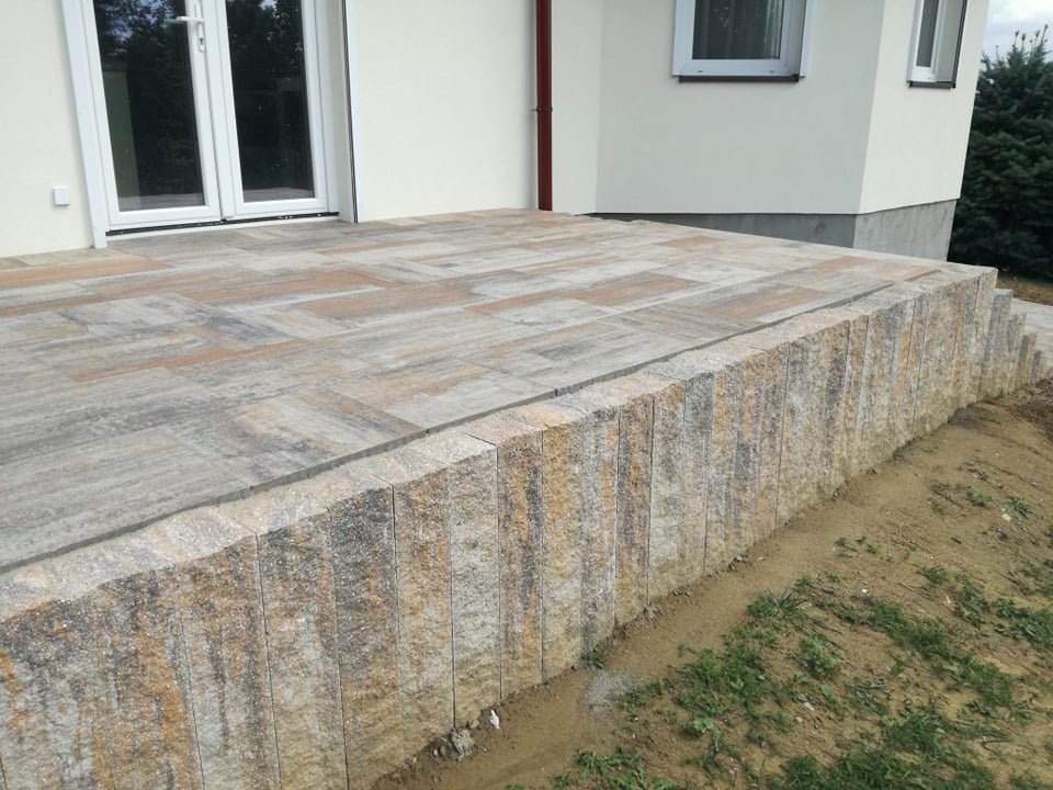 Podwyższony taras wyłożony wielobarwną kostką brukową, widok z boku na pionowe elementy obrzeża, z jasną elewacją budynku w tle.