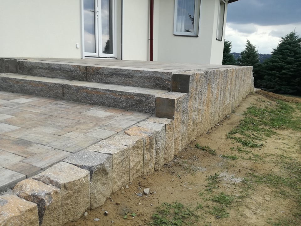 Schody wejściowe do domu wyłożone kostką brukową w odcieniach szarości i beżu, z pionową ścianką z kamienia o nieregularnej fakturze. Widoczne okna i fragment elewacji budynku w tle.