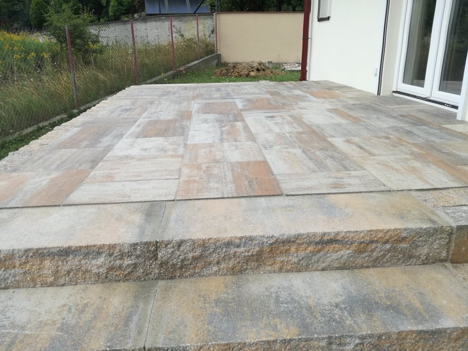 Ułożony taras z prostokątnych płyt brukowych w odcieniach beżu i szarości, z dwustopniowymi schodami z kostki, przylegający do białego budynku z podwójnymi drzwiami balkonowymi.