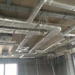 Surowy betonowy sufit z widocznymi, częściowo zaizolowanymi rurami wentylacyjnymi, metalowymi korytkami kablowymi i wiszącymi przewodami w stanie deweloperskim.