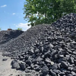 Dwa kopce czarnego węgla kamiennego na placu składowym, widoczne fragmenty ceglanego komina w tle, zieleń drzew i błękitne niebo.