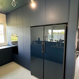 MYSTEK SPÓŁKA Z OGRANICZONĄ ODPOWIEDZIALNOŚCIĄ - Nowoczesna, granatowa kuchnia z zabudową po sufit i lodówką side-by-side. W tle okno i niedokończone oświetlenie sufitowe.