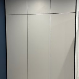 MYSTEK SPÓŁKA Z OGRANICZONĄ ODPOWIEDZIALNOŚCIĄ - Szafa wnękowa z szarymi, matowymi frontami i czarnymi obramowaniami, dopasowana do niebieskiej ściany w rogu pokoju, z widoczną podłogą imitującą drewno.