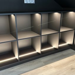 MYSTEK SPÓŁKA Z OGRANICZONĄ ODPOWIEDZIALNOŚCIĄ - Nowoczesna, grafitowa szafka z podświetleniem LED, wpasowana w skos poddasza, z widocznymi gniazdkami elektrycznymi na ścianie. Minimalistyczny design.