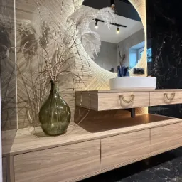 Nowoczesna łazienka z drewnianą szafką pod umywalkę, okrągłym lustrem z podświetleniem LED i marmurową podłogą. Wazon z trawami pampasowymi dodaje naturalnego akcentu.