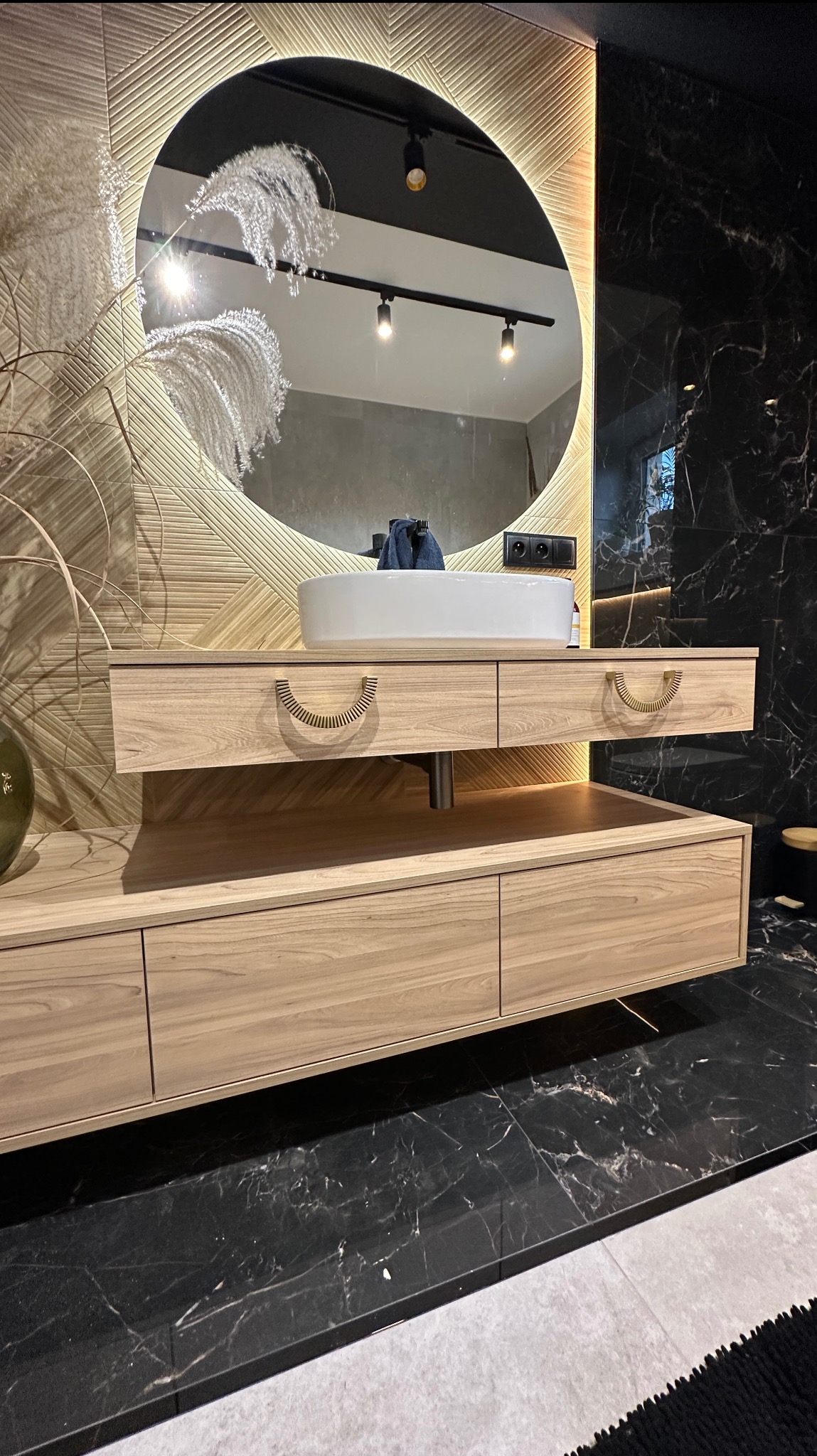 Nowoczesna łazienka z drewnianą szafką pod umywalkę, okrągłym lustrem z podświetleniem LED i marmurową ścianą.  Wyjątkowe uchwyty szuflad.