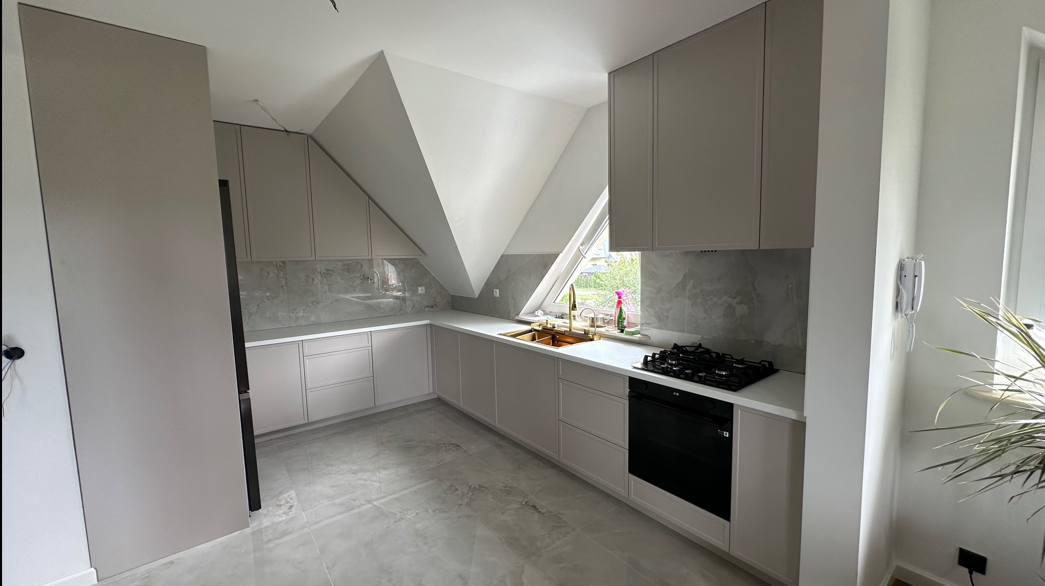 Jasna kuchnia na poddaszu ze złotą armaturą. Zabudowa pod skosem, marmurowe płytki nad blatem, płyta gazowa i piekarnik w zabudowie. Nowoczesny design.