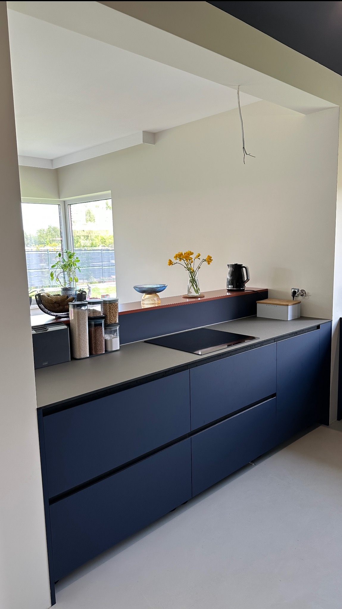Nowoczesna, granatowa kuchnia z szarym blatem, widok na okno i dekoracje. Minimalistyczny design z kwiatami i pojemnikami na blacie. Widoczne zawieszone kable.