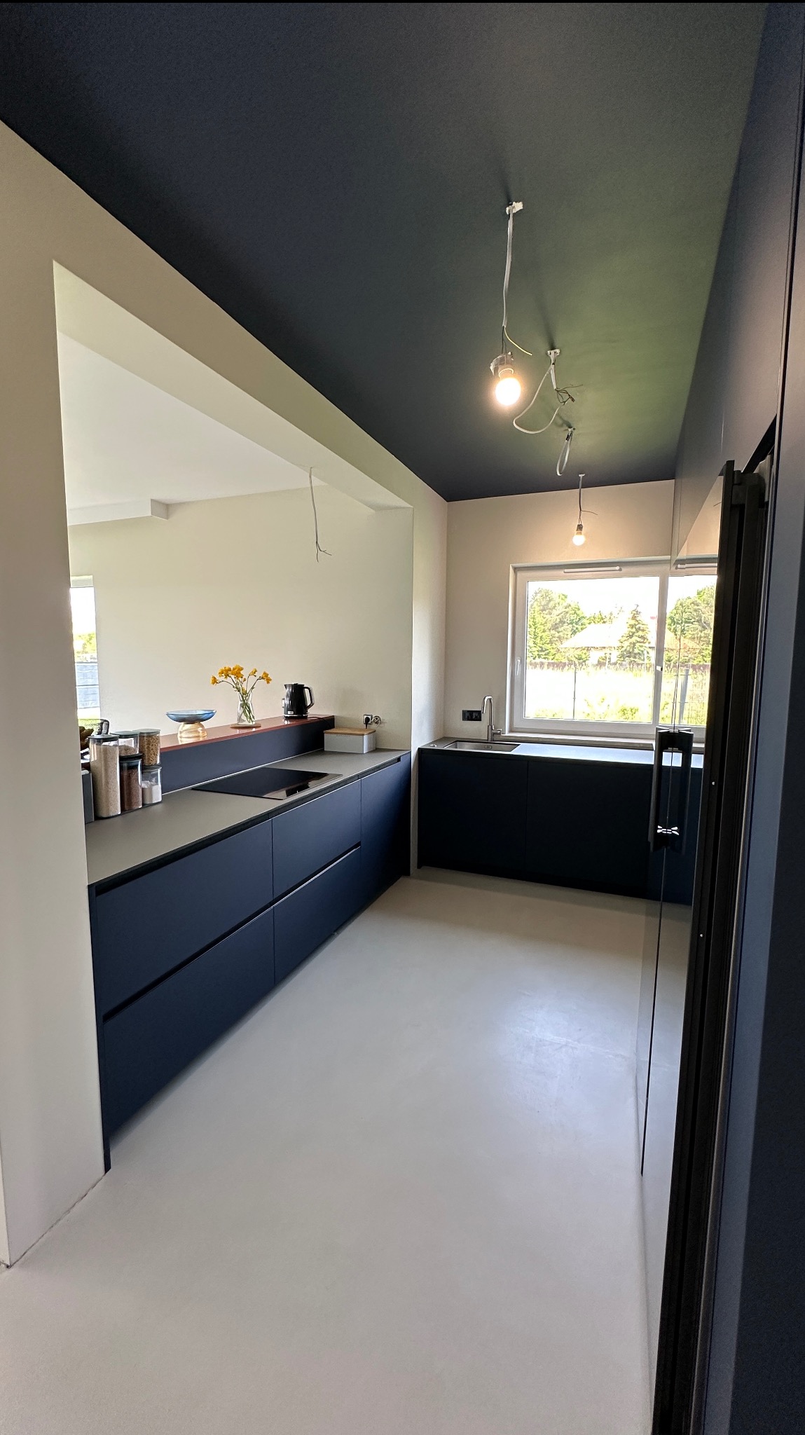 Nowoczesna kuchnia z ciemnymi szafkami i szarym blatem, z widokiem na okno i otwartą przestrzeń. Minimalistyczny design, sufit w kolorze granatowym. W tle kwiaty w wazonie.