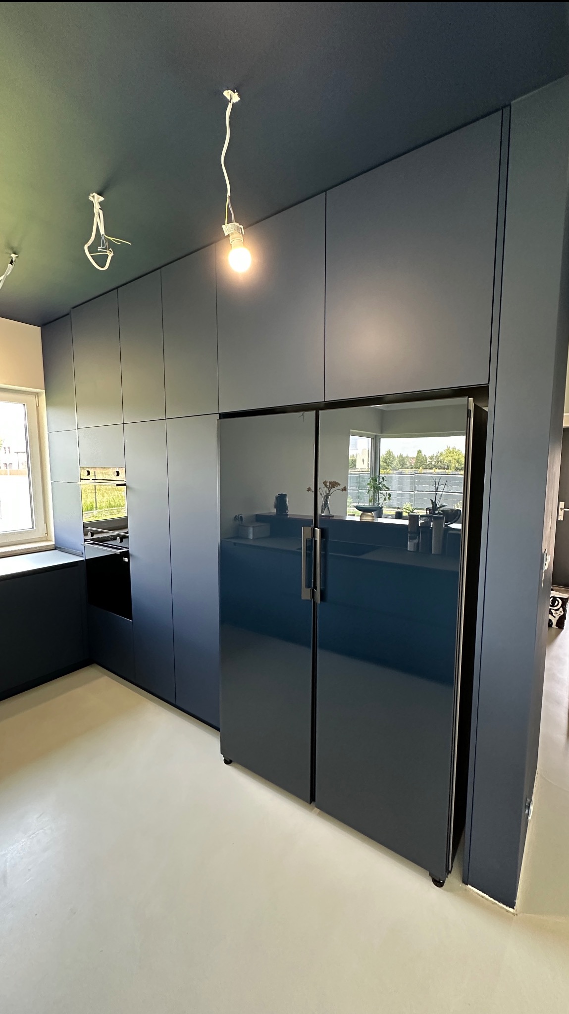 Nowoczesna, granatowa kuchnia z zabudową po sufit i lodówką side-by-side. W tle okno i niedokończone oświetlenie sufitowe.