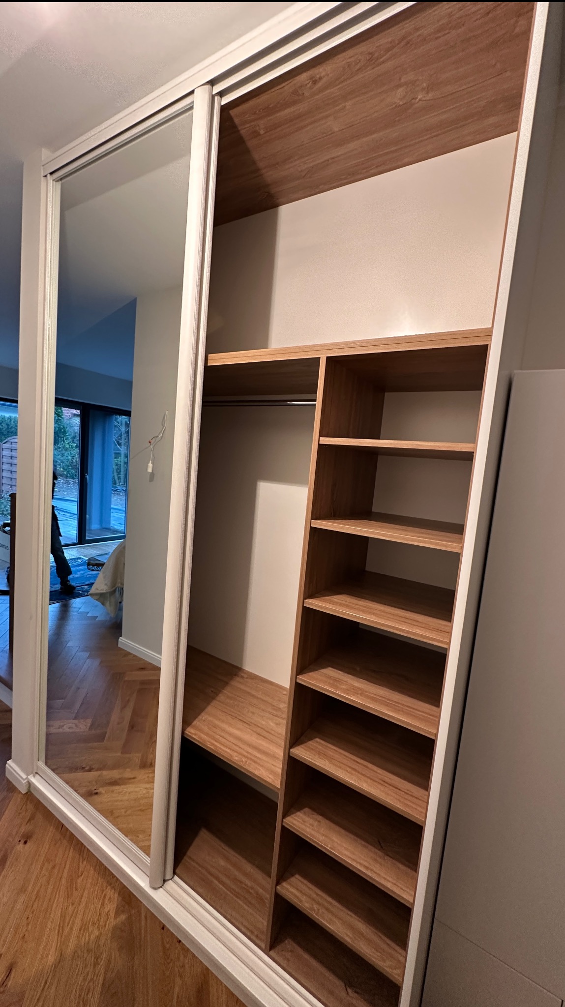 Wnętrze szafy wnękowej z lustrzanymi drzwiami przesuwnymi i drewnianymi półkami, widoczne z perspektywy wnętrza garderoby. Uwagę przykuwa minimalistyczny design i dbałość o detale.