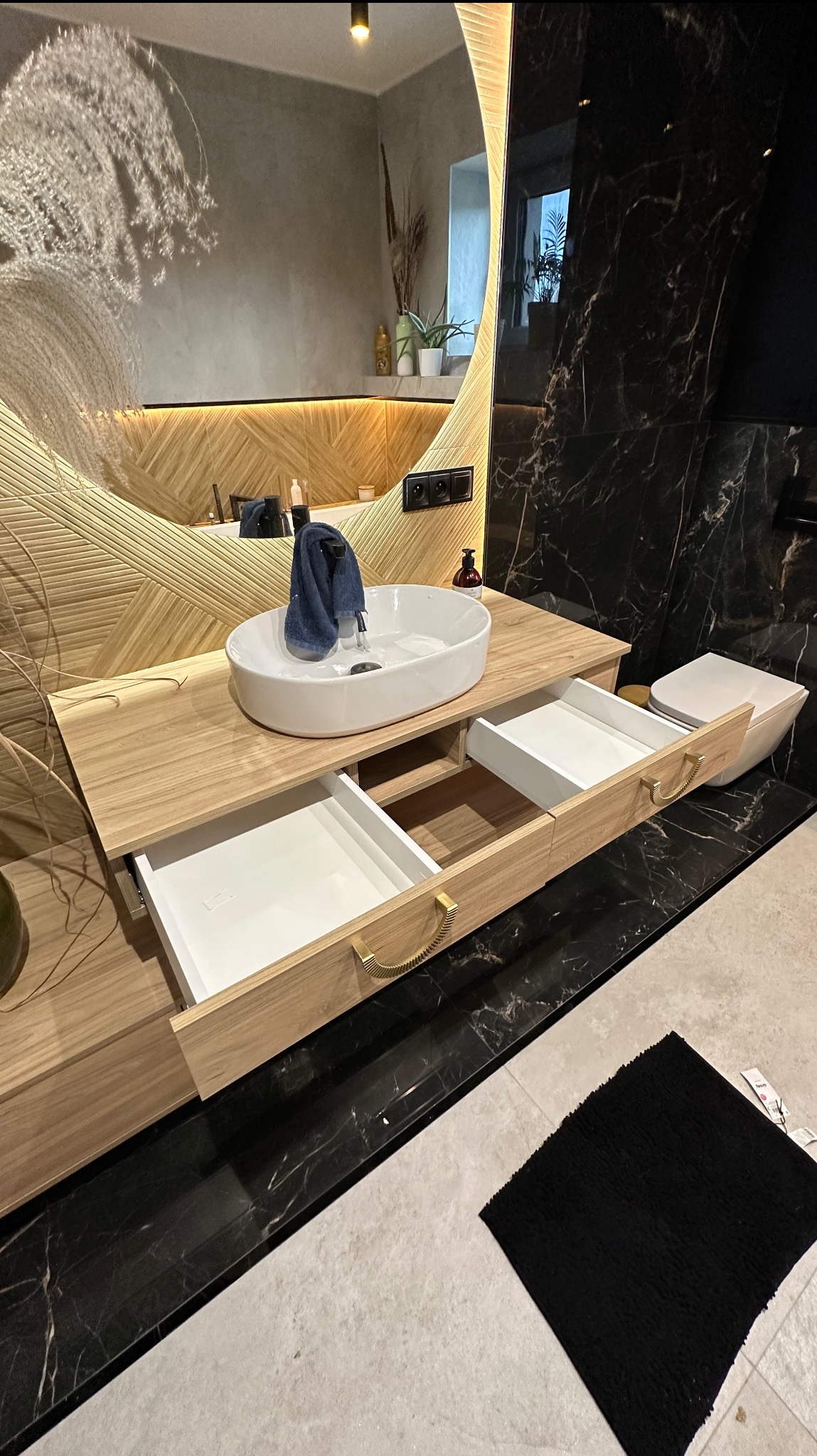 Nowoczesna szafka łazienkowa z owalną umywalką i otwartymi szufladami. Drewno o jasnym odcieniu, dekoracyjne uchwyty. W tle lustro z podświetleniem i marmurowe wykończenie.