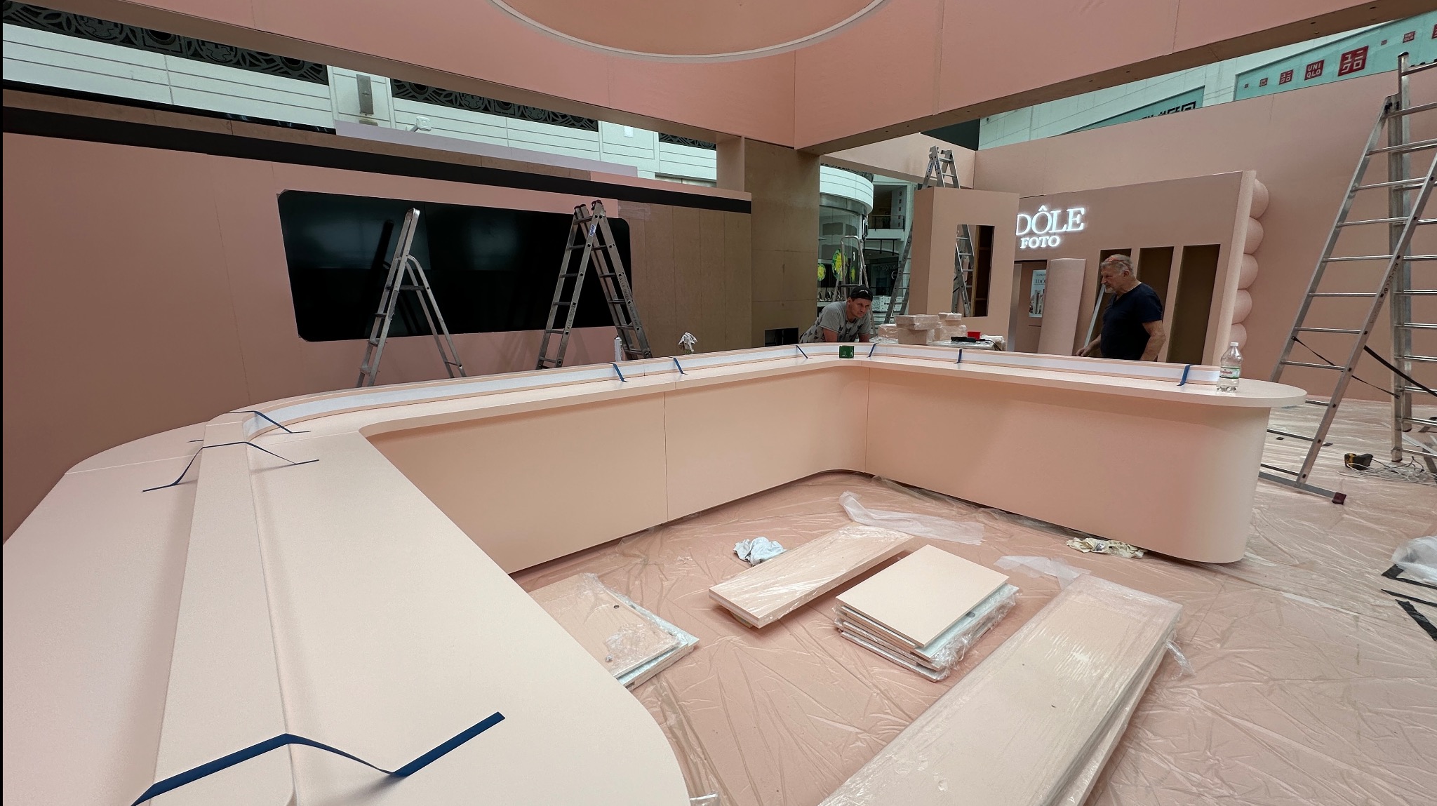 Montaż różowej lady recepcyjnej z logo 'Dole Foto' w przestrzeni handlowej. Widoczne elementy konstrukcyjne, drabiny i pracownicy podczas instalacji.