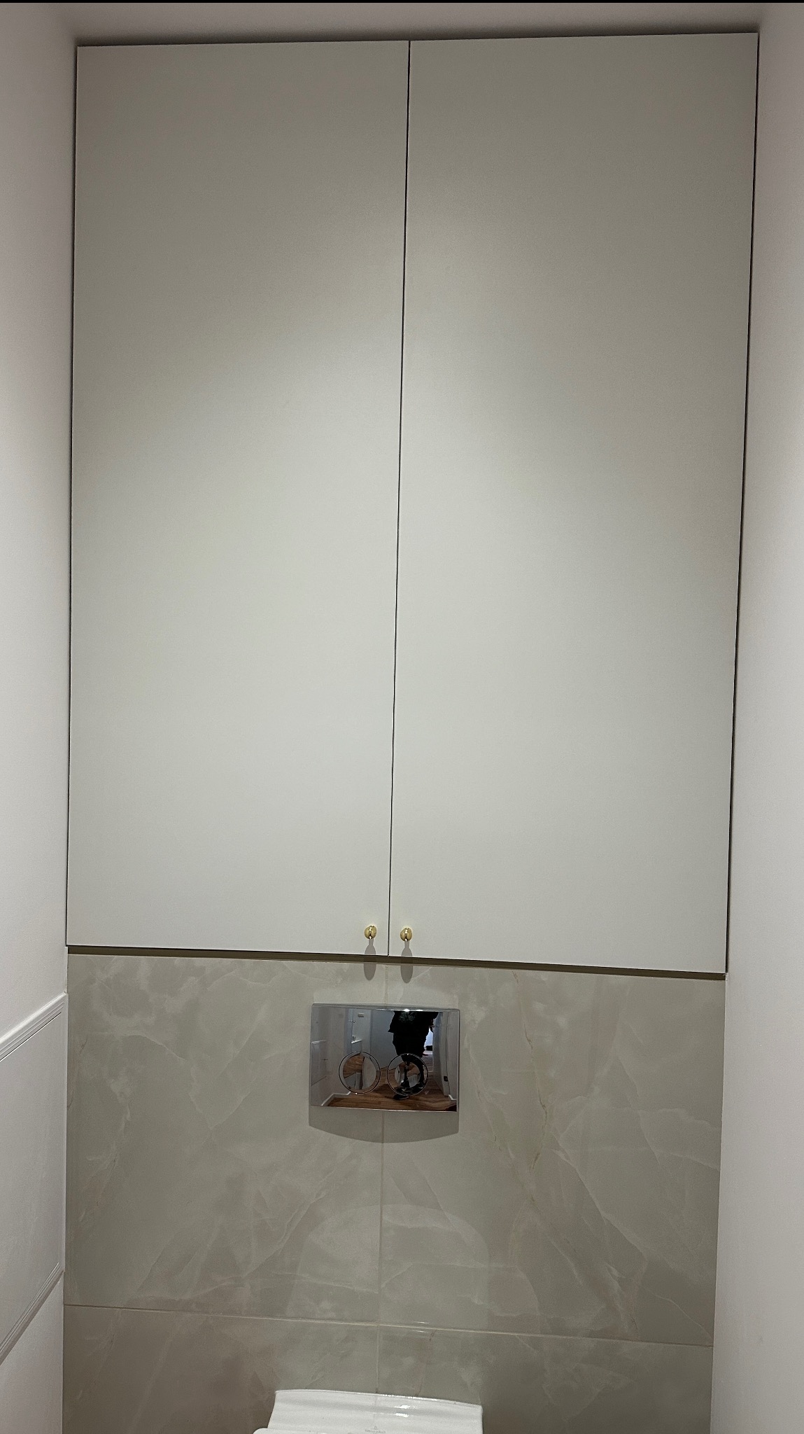 Biała szafka wisząca nad stelażem WC z marmurowymi płytkami. Złote gałki dodają elegancji. Minimalistyczny design w nowoczesnej łazience.