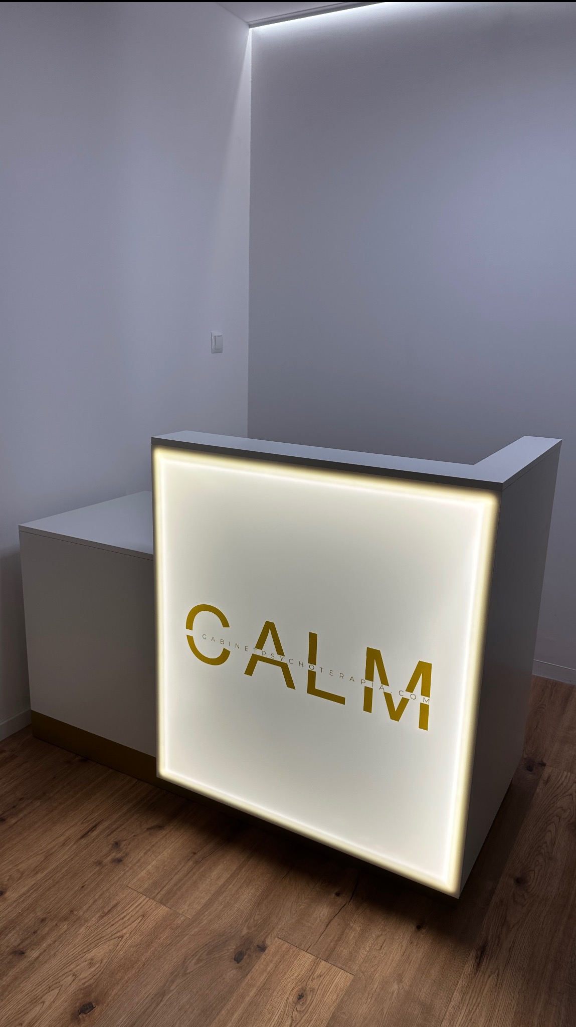 Biała lada recepcyjna z podświetlanym logo 'CALM Gabinet Psychoterapii' na drewnianej podłodze, w minimalistycznym wnętrzu z oświetleniem LED.