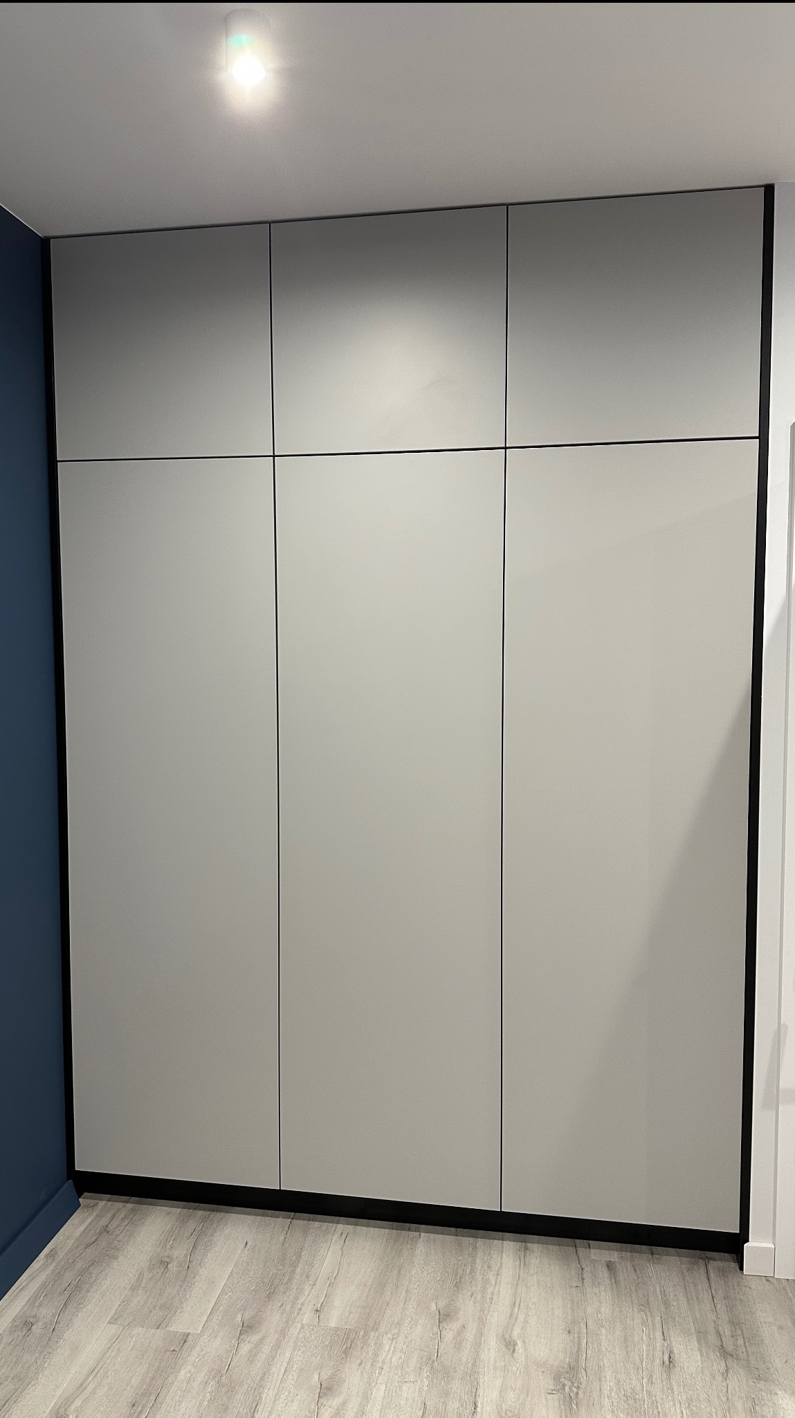 Szafa wnękowa z szarymi, matowymi frontami i czarnymi obramowaniami, dopasowana do niebieskiej ściany w rogu pokoju, z widoczną podłogą imitującą drewno.