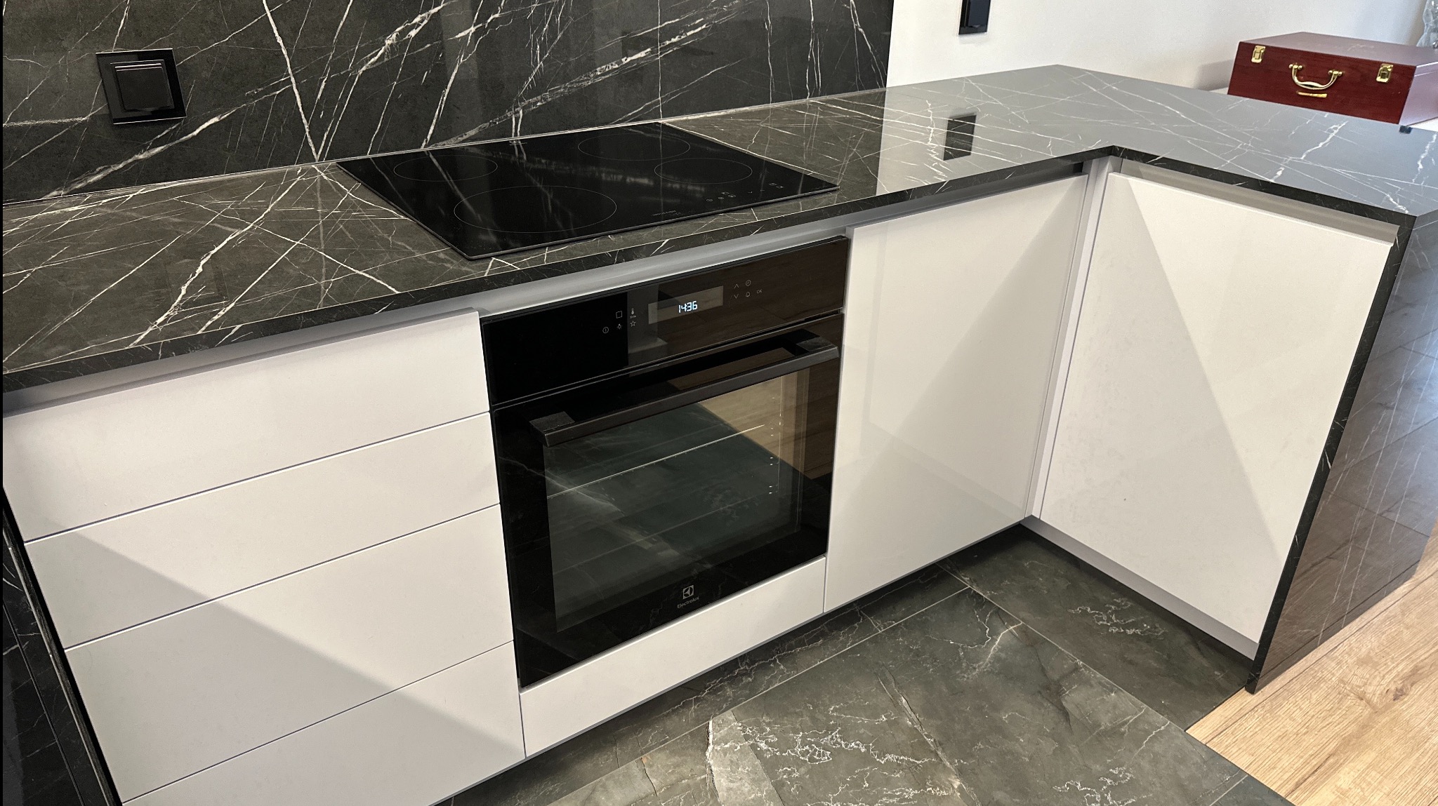 Nowoczesna kuchnia: białe fronty szafek, czarny blat z marmurowym wzorem, płyta indukcyjna i piekarnik Electrolux. W tle dekoracyjna skrzynia.