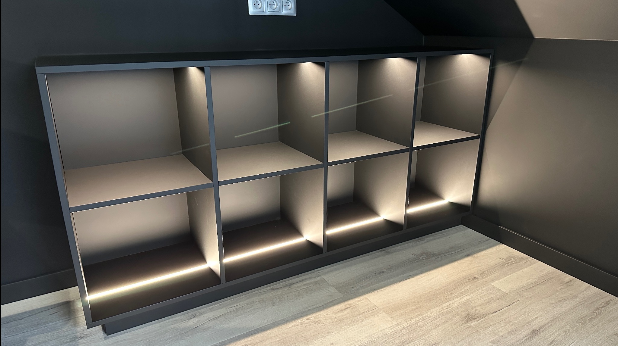Nowoczesna, grafitowa szafka z podświetleniem LED, wpasowana w skos poddasza, z widocznymi gniazdkami elektrycznymi na ścianie. Minimalistyczny design.