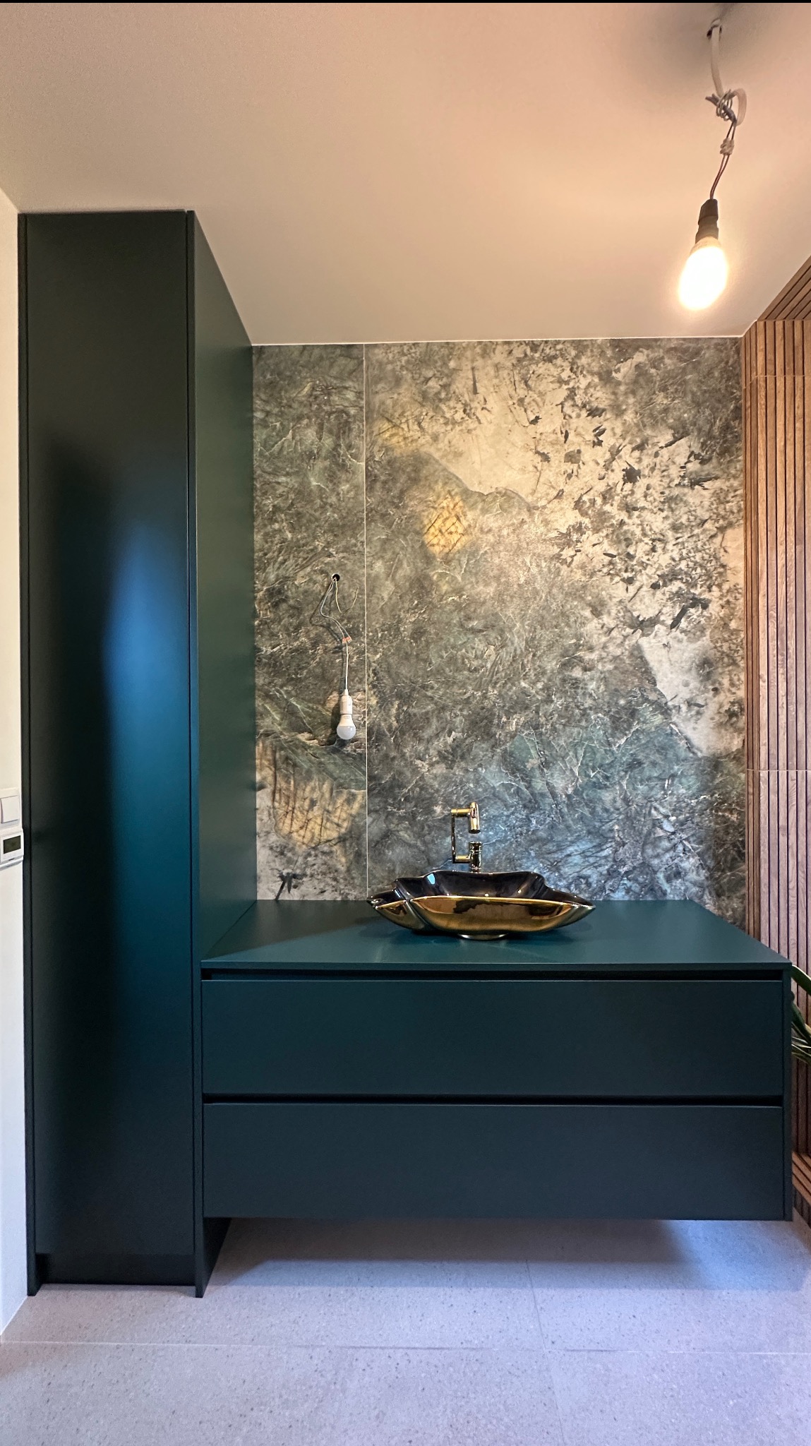 Nowoczesna łazienka z zielonymi szafkami, złotą umywalką i marmurową ścianą. W tle widoczna żarówka bez klosza i drewniane panele ścienne.