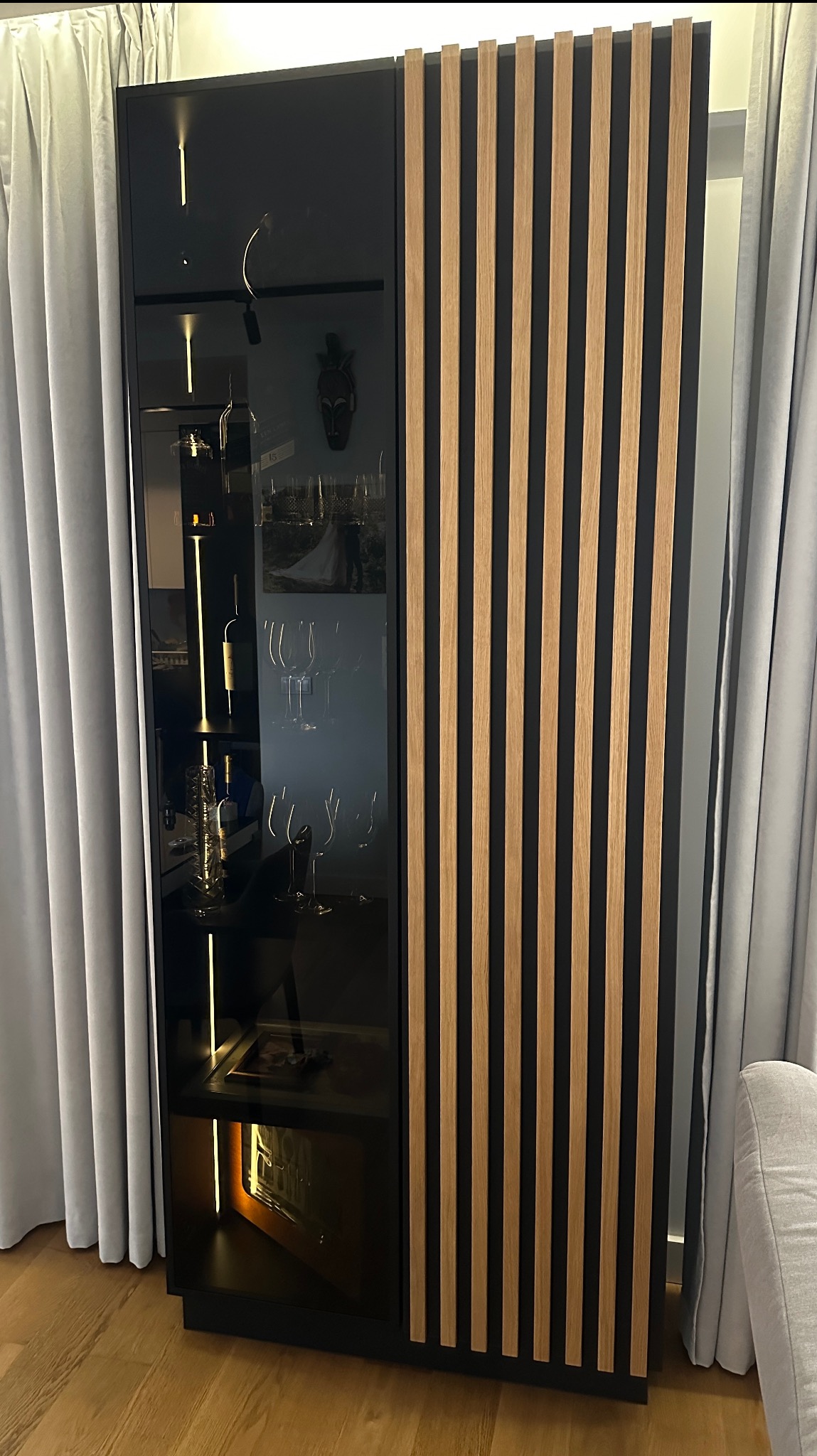Nowoczesna szafa na wymiar z czarnym frontem i drewnianymi lamelami, podświetlana wewnątrz, stojąca na drewnianej podłodze, obok zasłony.