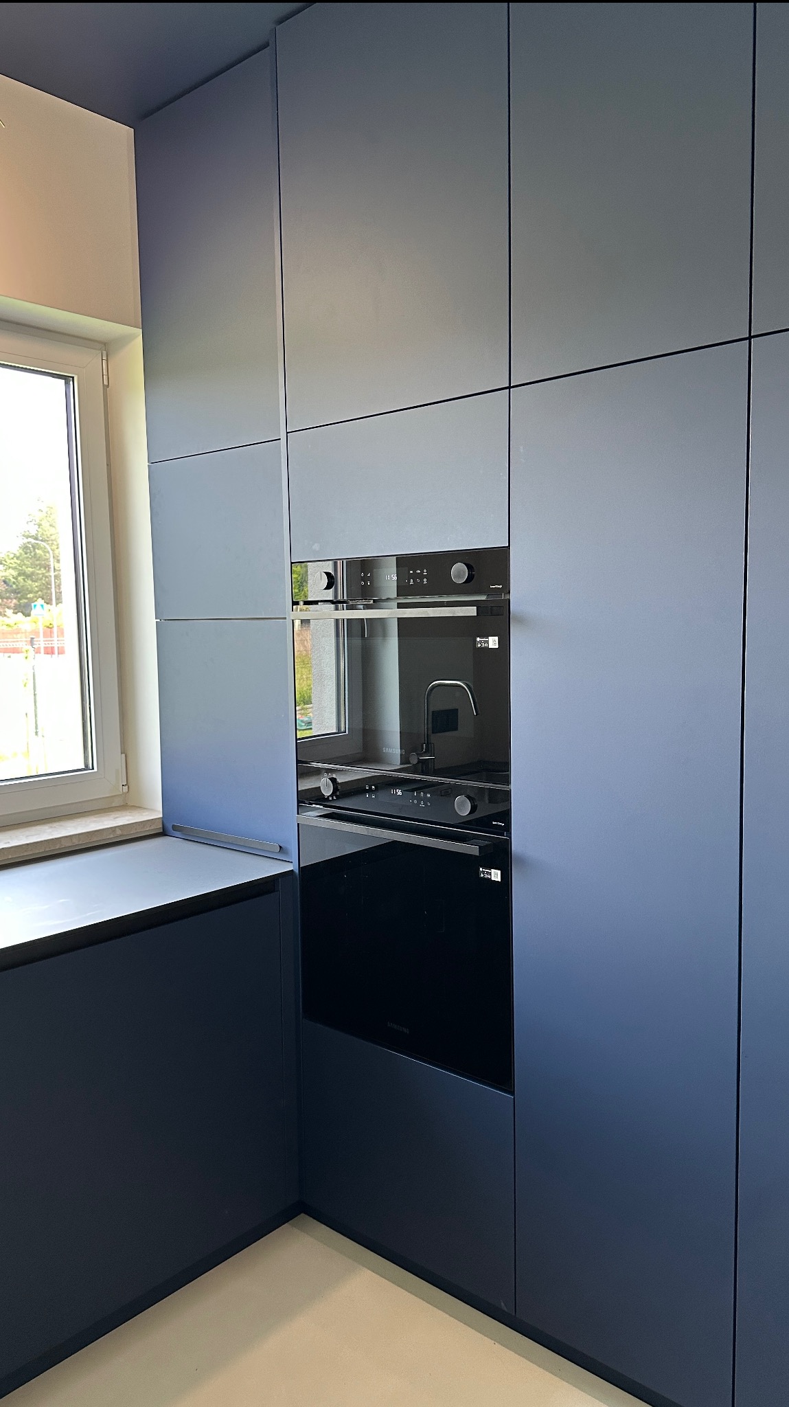 Nowoczesna, grafitowa zabudowa kuchenna z piekarnikiem i płytą w minimalistycznym stylu. Elementy bezuchwytowe, gładkie fronty, widok na okno i zieleń na zewnątrz.