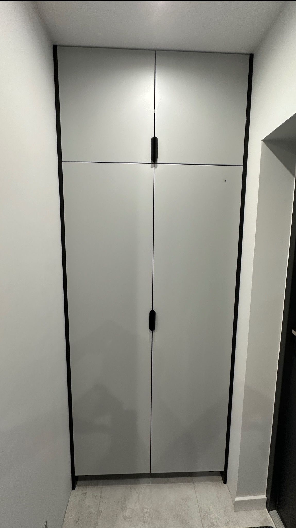 Jasnoszara szafa wnękowa z czarnymi detalami i minimalistycznymi uchwytami, wkomponowana w jasne wnętrze, widoczna z perspektywy podłogi.